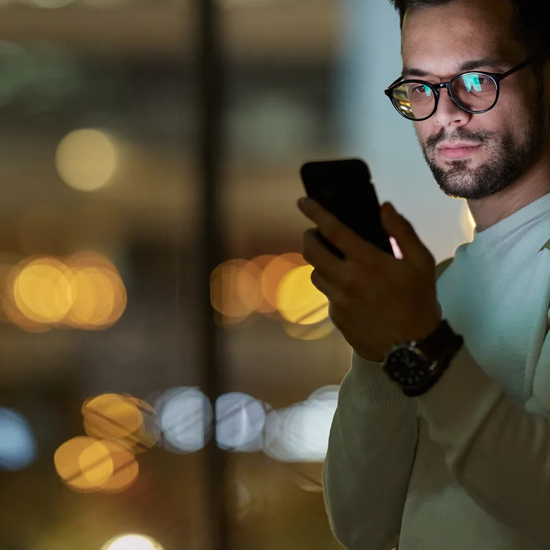 Library photo: a man checking his phone against city lights at night